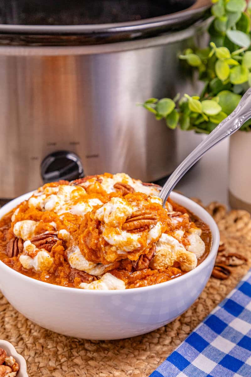 A white bowl full of sweet potato casserole topped with marshmallows and pecans with a slow cooker seen in the background.