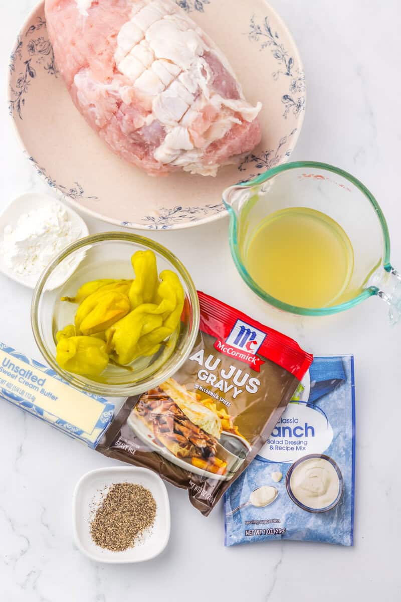 A raw turkey breast, chicken broth, pepperonicini peppers, butter, and seasoning mixes on a white background.