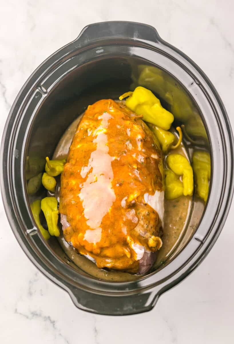 A turkey breat in a slow cooker covered in Mississippi butter and surrounded by pepperoncini peppers.