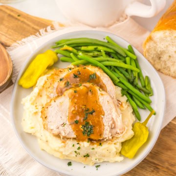 Crockpot Mississippi turkey breast, sliced and topped with gravy on a white plate with mashed potatoes, green beans, and pepperoncini peppers.