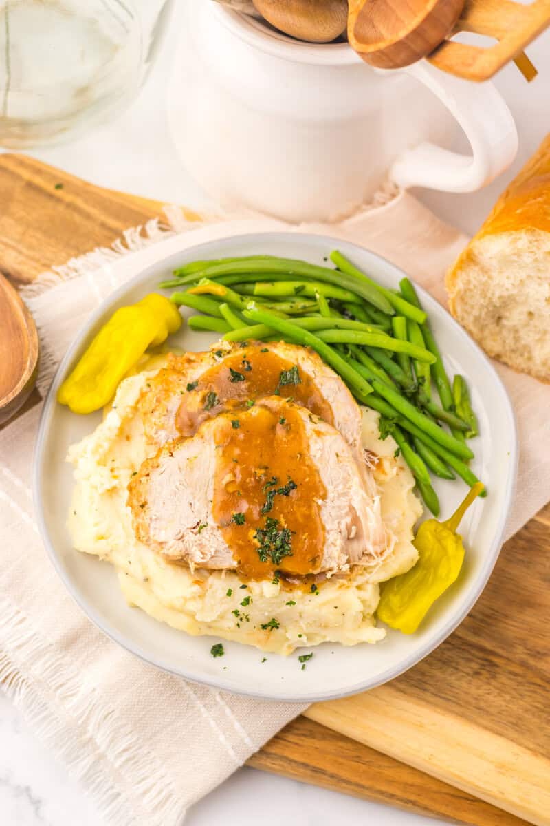 Crockpot Mississippi turkey breast, sliced and topped with gravy on a white plate with mashed potatoes, green beans, and pepperoncini peppers. 