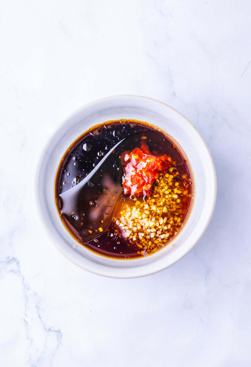 A white bowl with soy sauce, minced garlic, sesame seeds, and a dollop of red chili paste on a white marble surface.