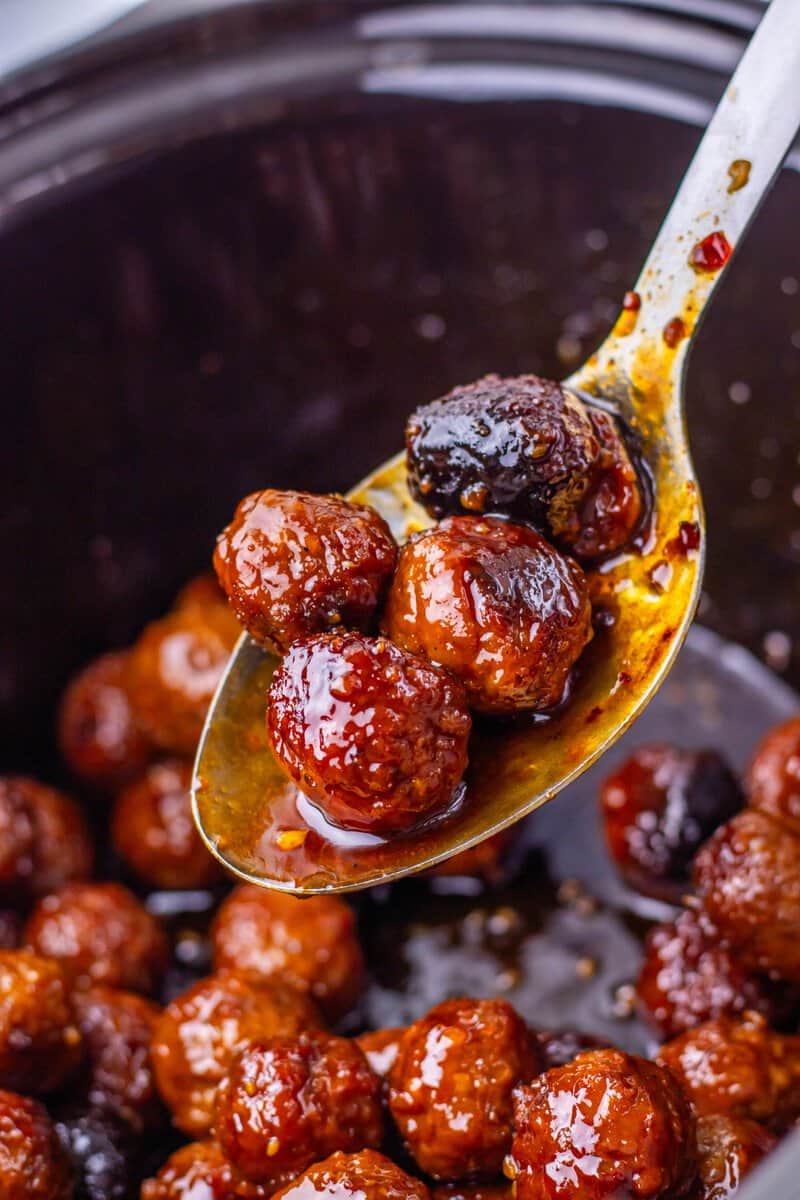 A spoon holding glazed meatballs over a slow cooker filled with more glazed meatballs in sauce.