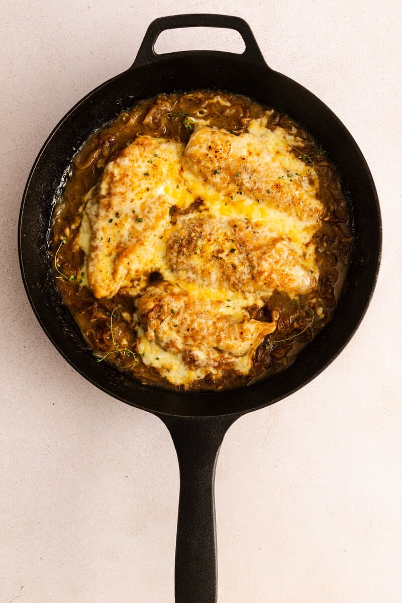 French onion chicken topped with melted cheese in a black cast iron skillet on a light background.