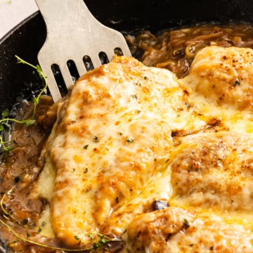 A piece of chicken being lifted out of a cast iron skillet full of French Onion Chicken Bake with a metal spatula.
