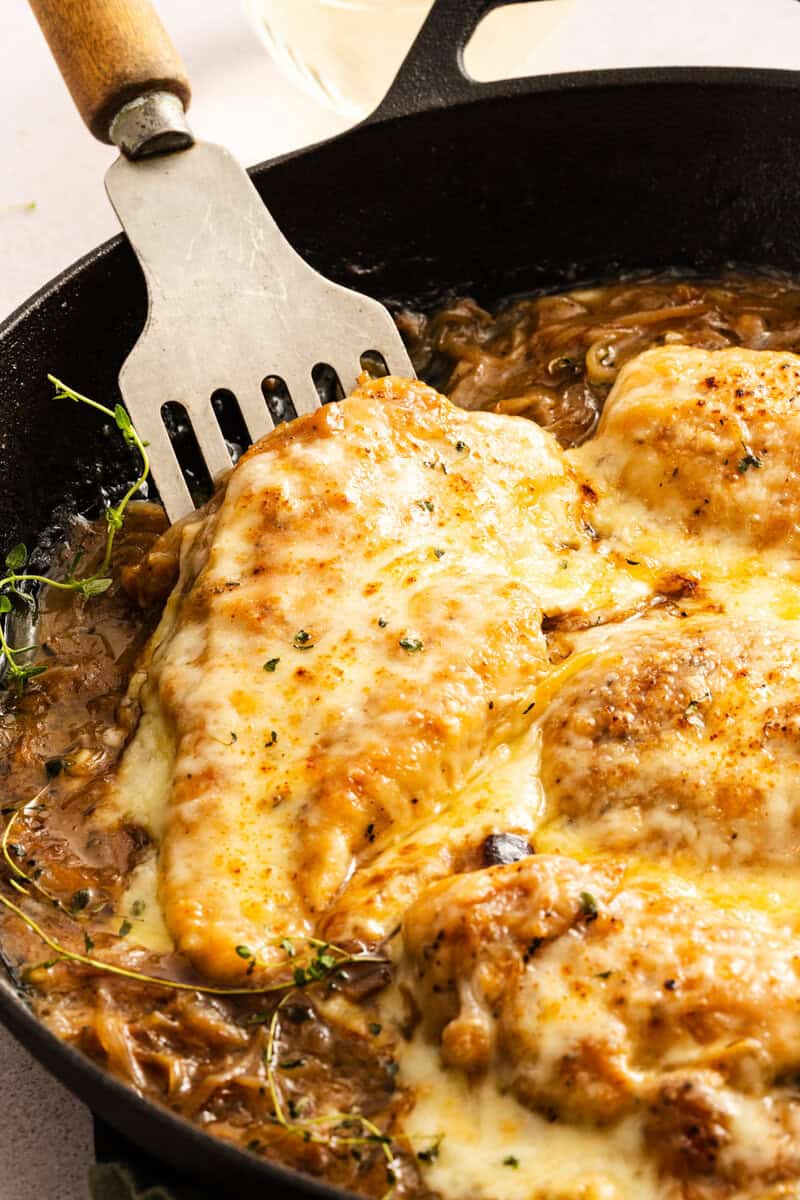 A piece of chicken being lifted out of a cast iron skillet full of French Onion Chicken Bake with a metal spatula. 