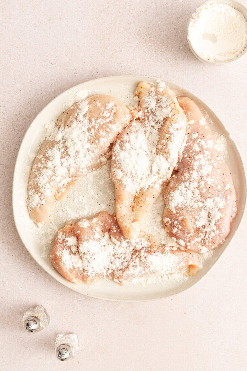 Raw chicken breast on a white plate, sprinkled with seasoning and flour.
