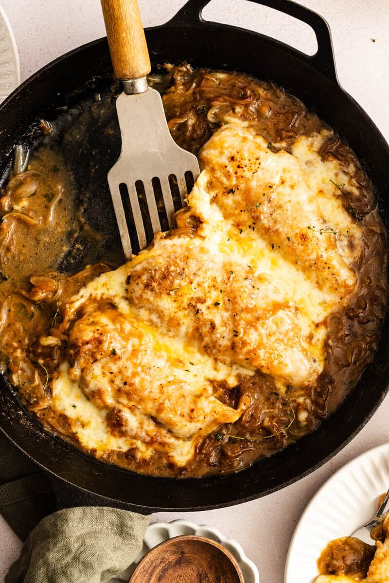 A spatula lifting a chicken breast out of a skillet of French onion chicken.