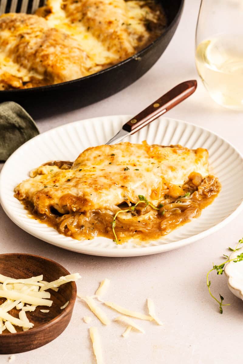 A serving of French onion chicken bake on a white plate with a fork with the skillet seen in the background.