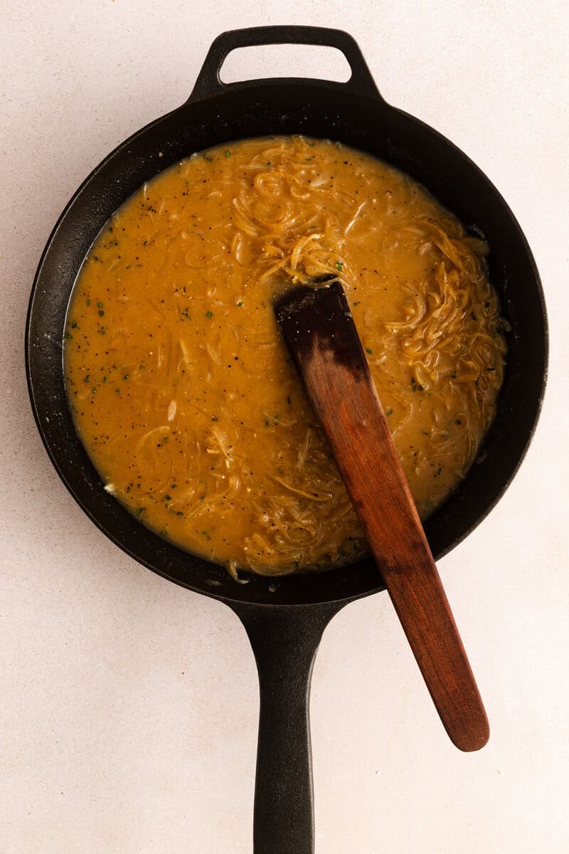Caramelized onions in a brown sauce in a black cast iron skillet with a wooden spatula.