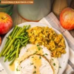 A plate of slow cooker turkey breast with gravy, green beans, stuffing, and mashed potatoes is displayed on a table with apples, a pot, and a towel.